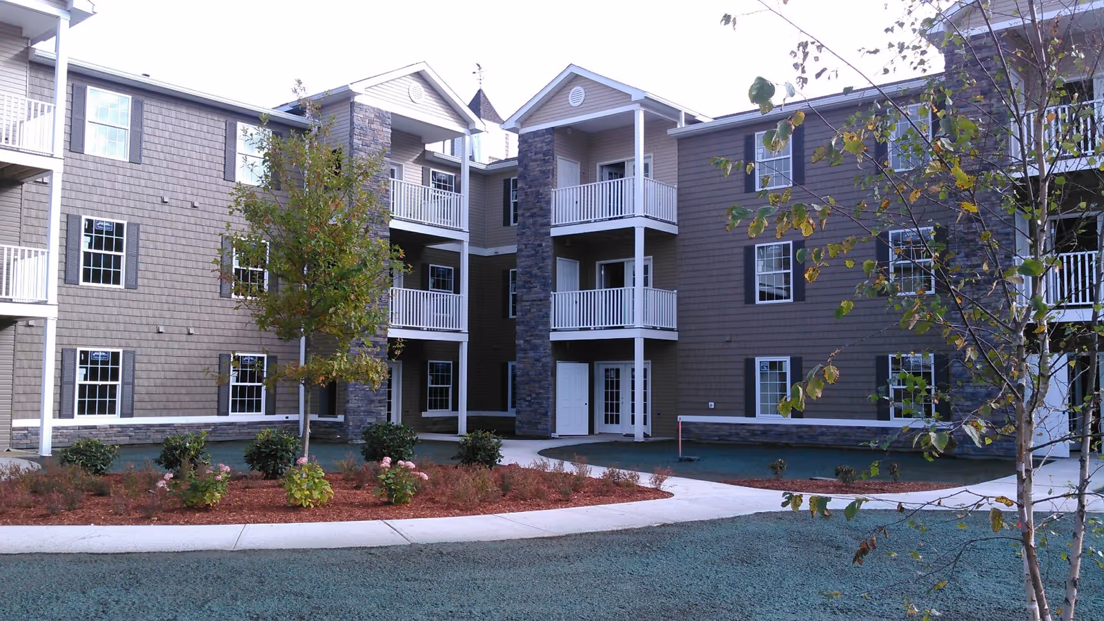 Exterior view of a multi-story residential building with balconies, surrounded by landscaped grounds including trees, shrubs, and a curved sidewalk.