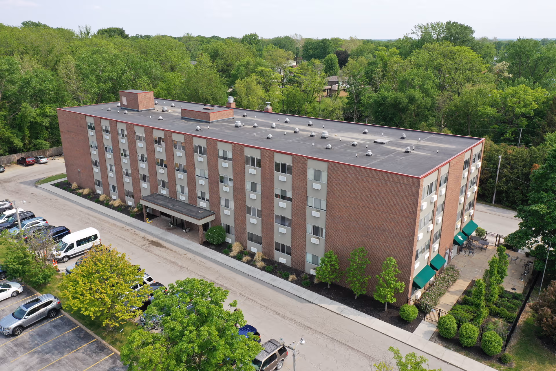 Aerial view of a four-story brick building with multiple windows, surrounded by trees and greenery. There is a parking lot with several cars and a white van parked near the building. The building has a covered entrance and green awnings on one side with outdoor seating visible. The area is lush with trees and vegetation.