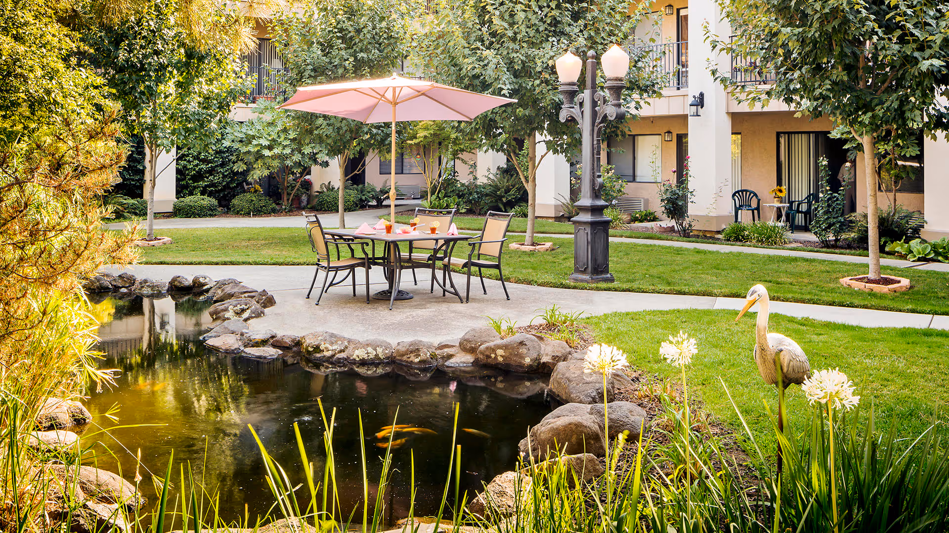 Outdoor courtyard area with a small pond surrounded by rocks and plants, a table with four chairs under a pink umbrella, and a decorative bird statue near the pond. The background shows a building with balconies and greenery.