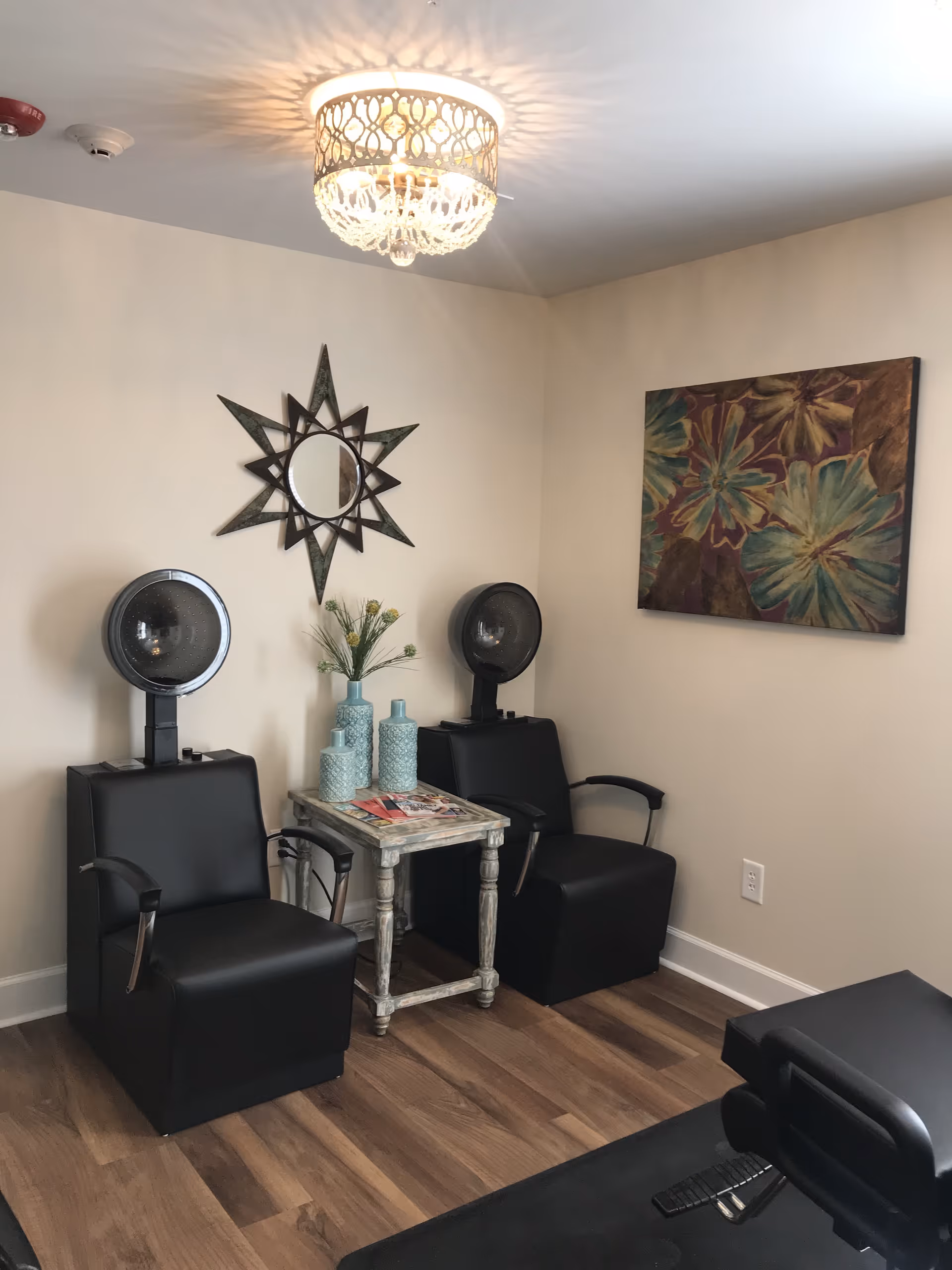 Interior room with two black salon hair dryer chairs positioned against a beige wall. Between the chairs is a small rustic wooden table with decorative blue vases and magazines on top. Above the table is a star-shaped mirror, and to the right is a colorful floral painting. The room has wood flooring and a decorative ceiling light fixture.