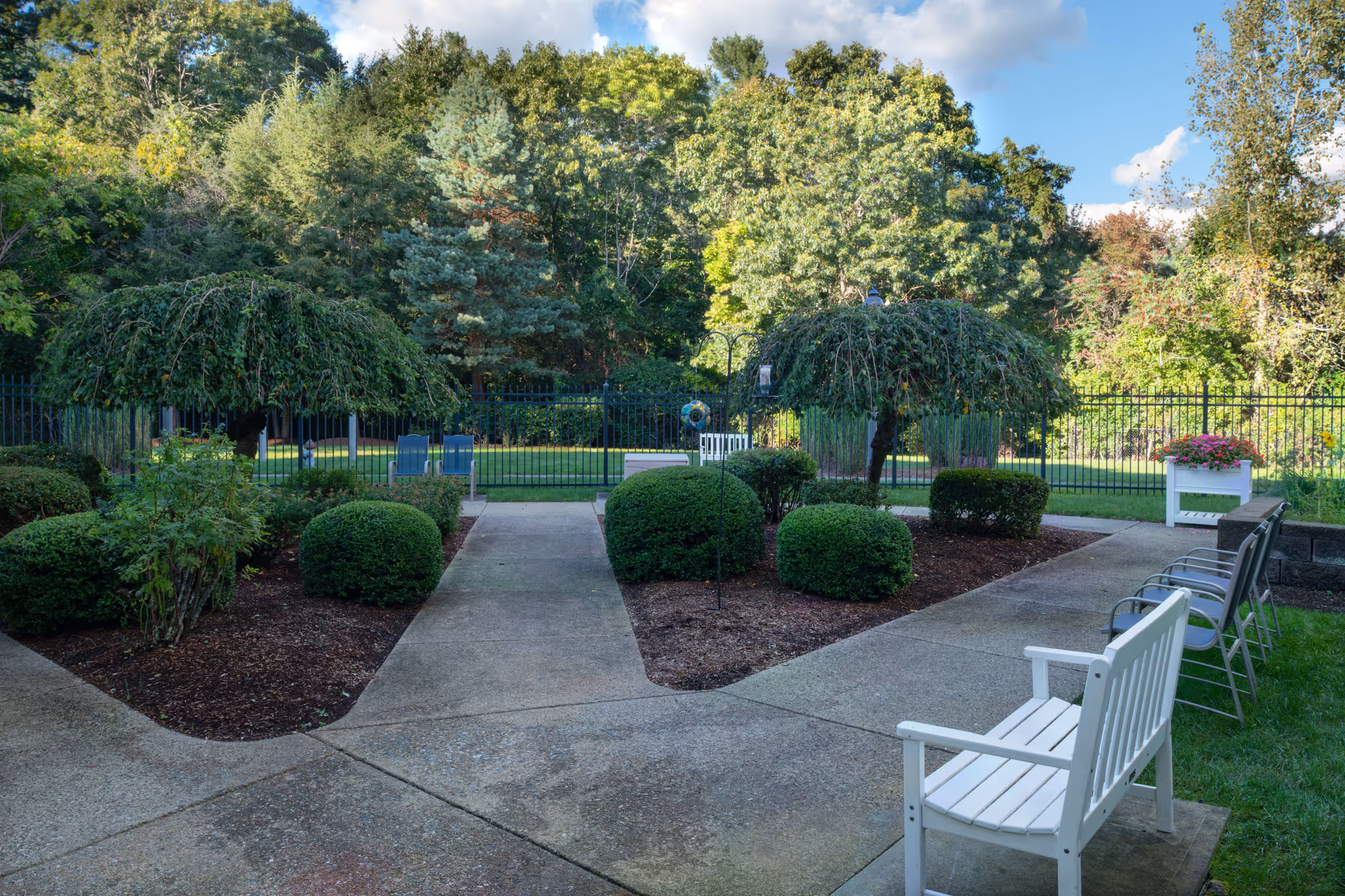 A peaceful outdoor garden area with a paved walkway, neatly trimmed bushes, two small weeping trees, several chairs, and a white bench. The garden is enclosed by a black metal fence with dense green trees in the background under a partly cloudy blue sky.