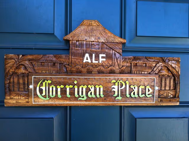 A carved wooden sign mounted on a dark blue door. The sign features a thatched roof design at the top with the letters 'ALF' and the name 'Corrigan Place' in stylized yellow-green lettering below.