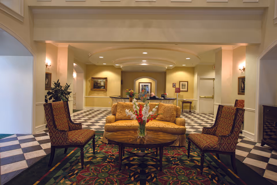 A cozy seating area in a senior living facility lobby with a patterned carpet, two upholstered chairs, a cushioned sofa, and a round wooden coffee table with a vase of flowers. The background shows a reception desk with a person seated behind it, framed artwork on the walls, and checkered black and white flooring.