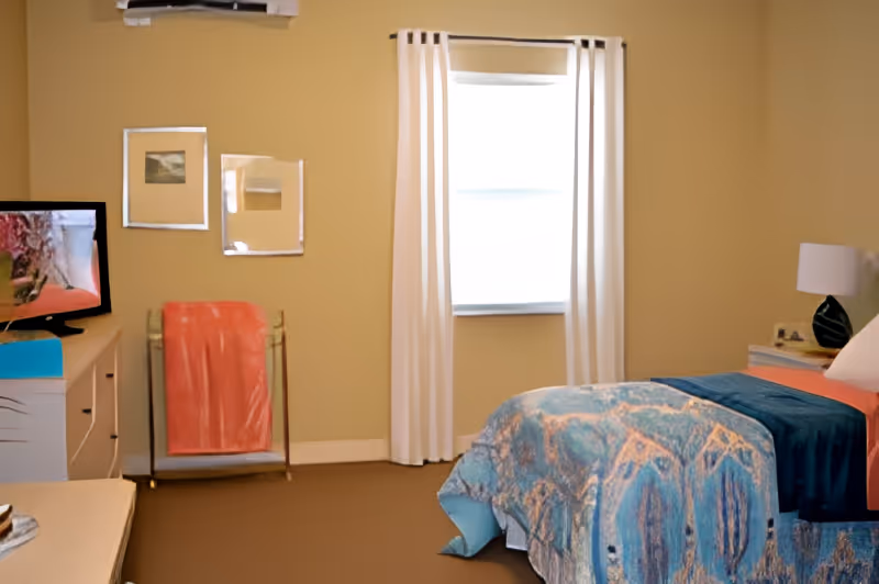 A cozy bedroom with a bed covered in a blue and orange patterned blanket, a bedside table with a lamp and books, a window with white curtains, a TV on a dresser, and a towel rack with an orange towel.