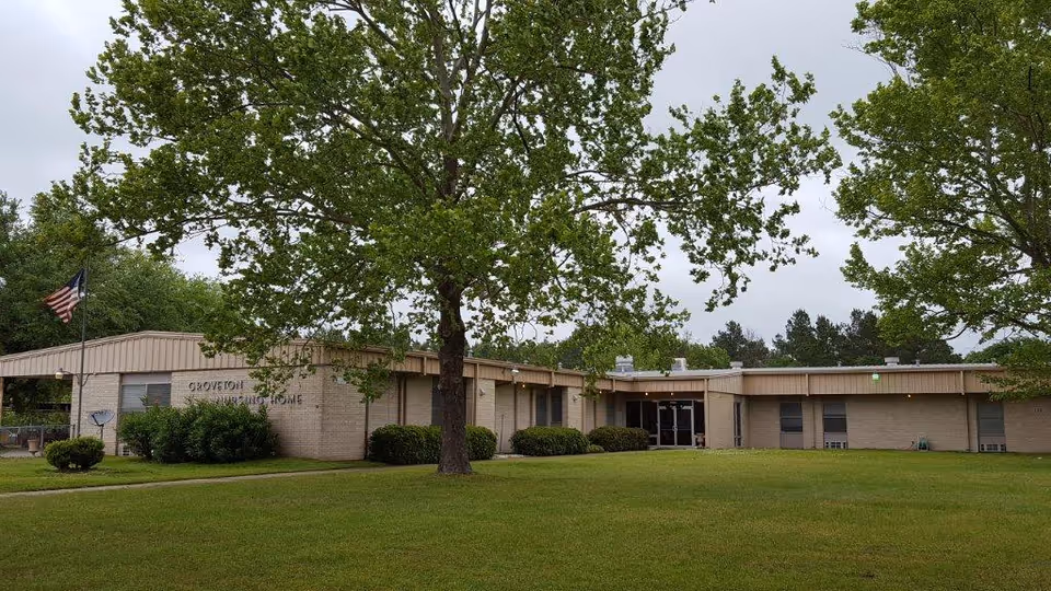 Exterior view of Groveton Nursing Home, a single-story beige brick building with a flat roof, surrounded by green grass and large trees. An American flag is visible on the left side near the entrance.