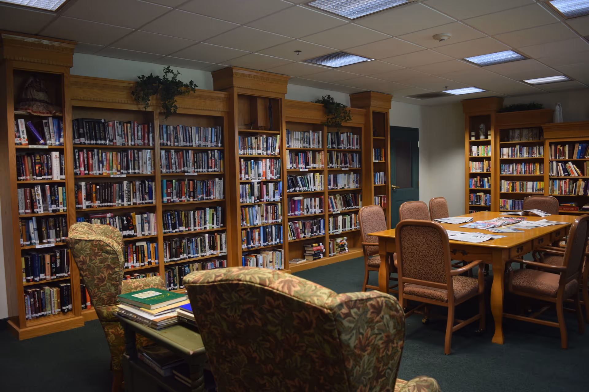 A cozy library room with wooden bookshelves filled with books along the walls. There is a wooden table in the center surrounded by six upholstered chairs. Two patterned armchairs are in the foreground, and some magazines and newspapers are spread out on the table. The room has a green carpet and a drop ceiling with fluorescent lights.