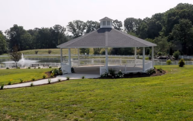 A white gazebo with a gray roof situated on a grassy area near a pond with a water fountain, surrounded by trees and a white fence in the background.