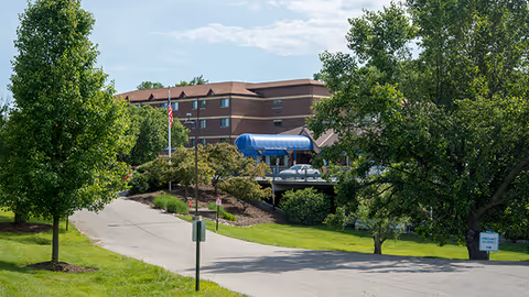 Exterior view of Western Hills Retirement Village showing a multi-story brick building with a blue awning over the entrance, surrounded by green trees and a paved driveway leading up to the entrance.