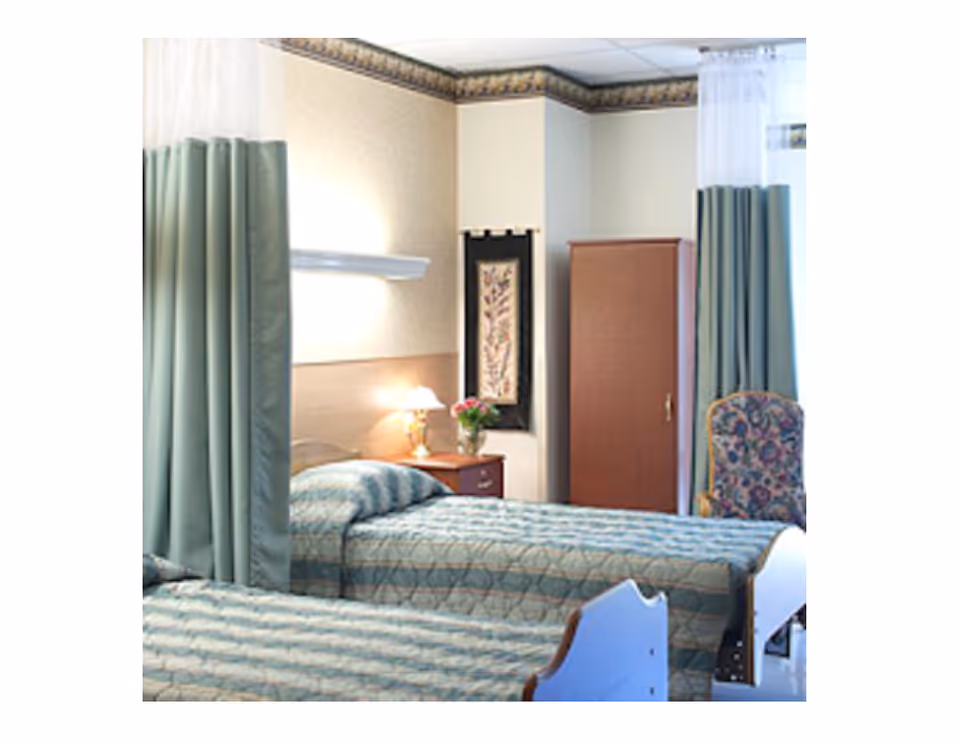 A room with two single beds covered in patterned bedspreads, separated by a green privacy curtain. There is a wooden nightstand with a lamp and a vase of flowers between the beds. A floral upholstered chair and a wooden wardrobe are visible in the background, along with a decorative wall hanging.