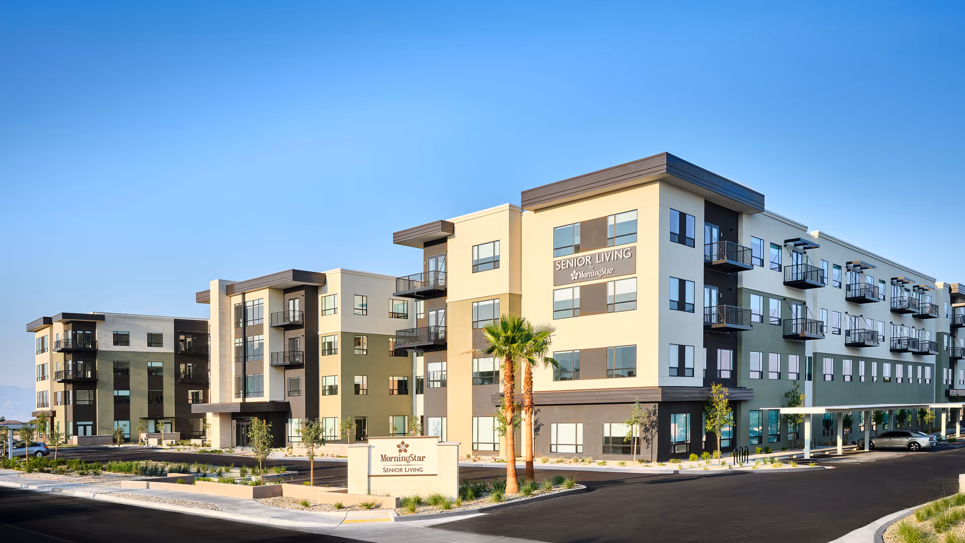 Exterior view of a modern four-story senior living facility building with multiple balconies, large windows, and a sign in front that reads 'MorningStar Senior Living'. The building is surrounded by a paved driveway, landscaping with palm trees, and clear blue sky overhead.