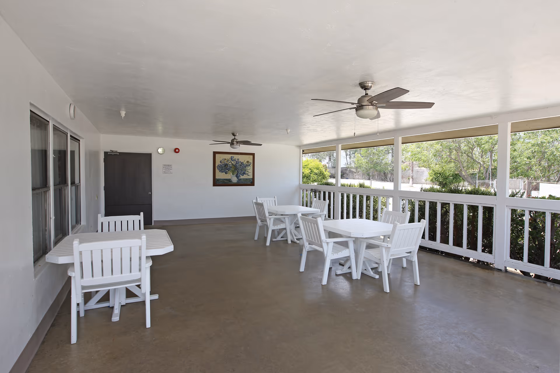 Covered outdoor patio area with white tables and chairs, ceiling fans, and a railing overlooking greenery outside. A framed picture hangs on the far wall next to a closed door.