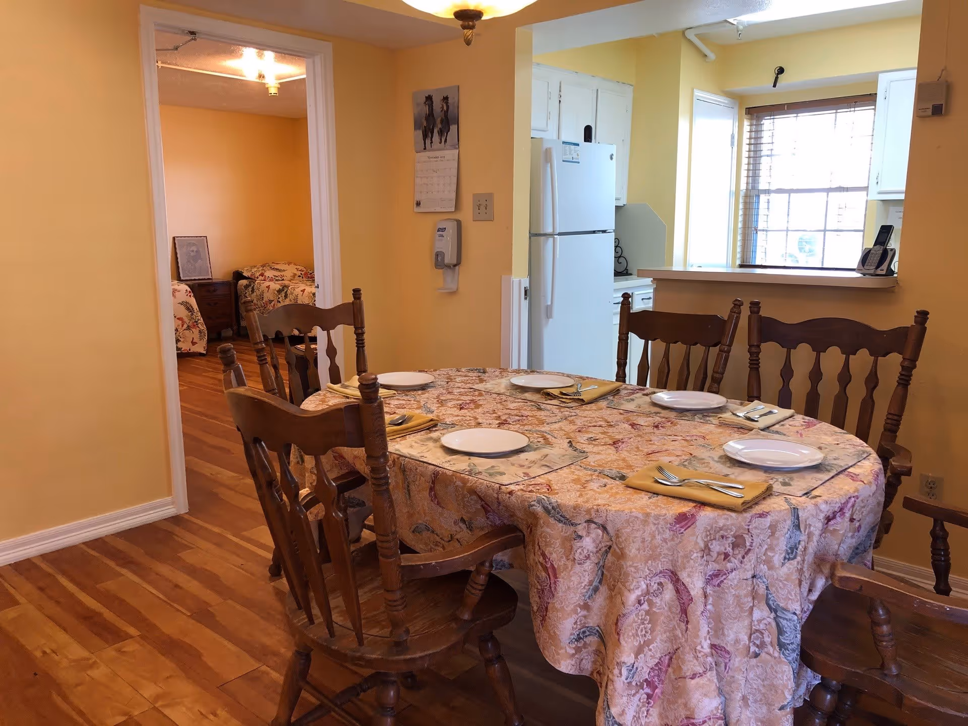 Dining area with a round table covered with a patterned tablecloth set with plates, napkins, and silverware. Six wooden chairs surround the table. In the background, there is a kitchen with white cabinets and a refrigerator, and an adjacent room with two beds and a nightstand.