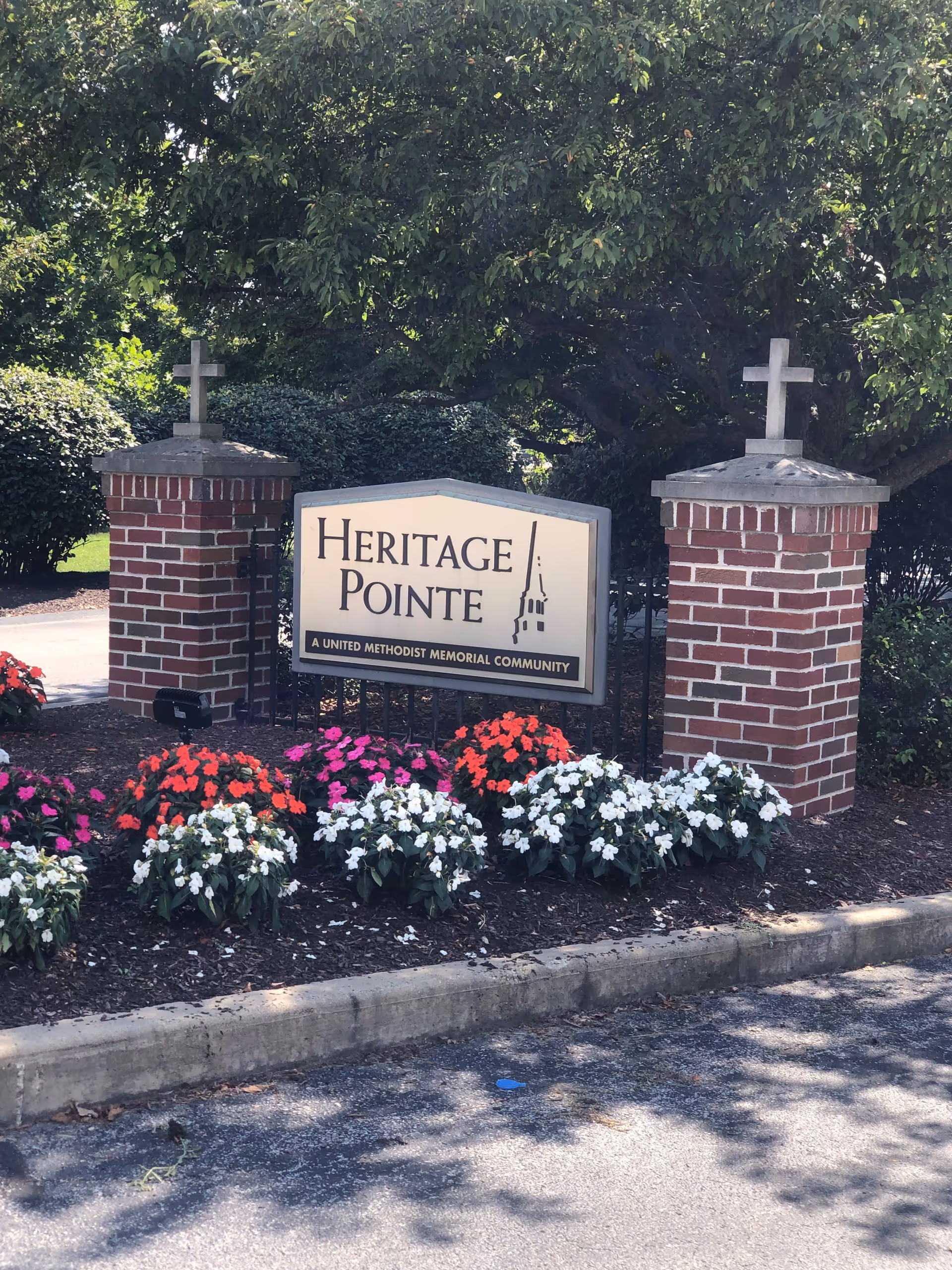 Entrance sign for Heritage Pointe, a United Methodist Memorial Community, flanked by two brick pillars topped with crosses, surrounded by colorful flowers and greenery.