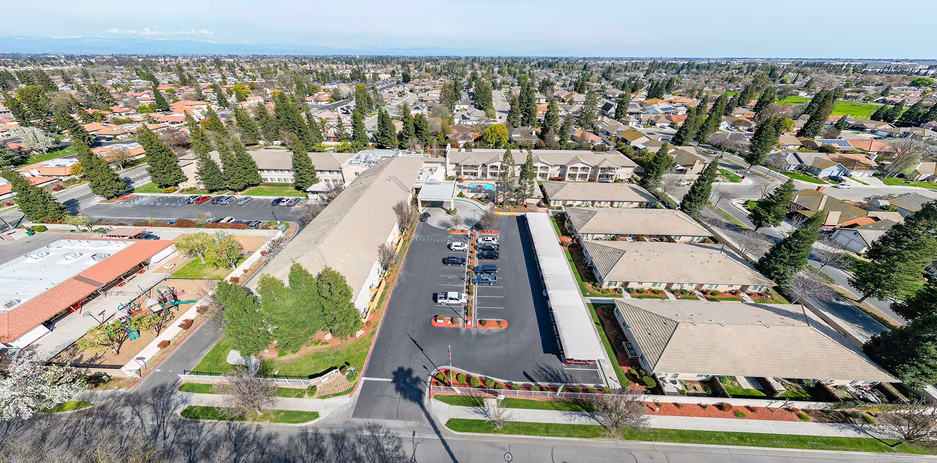 Aerial view of Cogir of Cedar Creek senior living facility showing multiple buildings with beige roofs, a parking lot with cars, landscaped areas with trees and bushes, a playground, and a swimming pool. Surrounding the facility are residential neighborhoods with houses and streets extending into the distance.