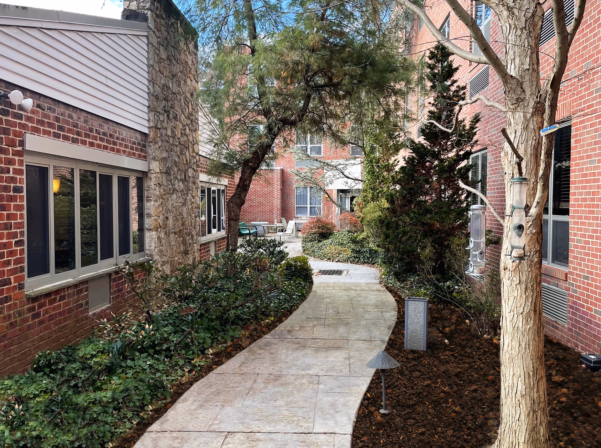 A paved walkway winding through a garden area between two brick buildings. The garden features various shrubs, small trees, and bird feeders hanging from a tree on the right. In the background, there are outdoor chairs and tables near the building entrance.
