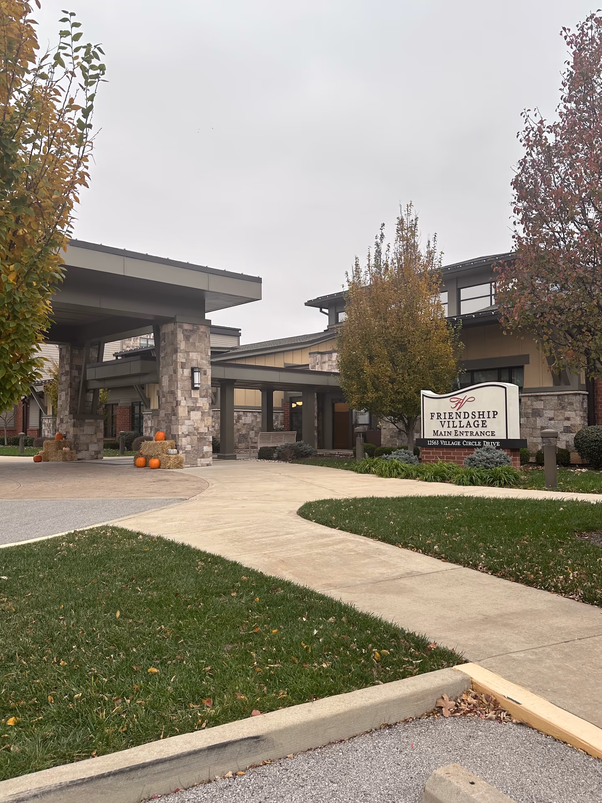 Exterior view of the main entrance to Friendship Village Sunset Hills, showing a covered driveway with stone pillars, a paved walkway, green lawns, autumn trees, and a sign with the facility name and address. Several pumpkins and hay bales are placed near the entrance as seasonal decorations.