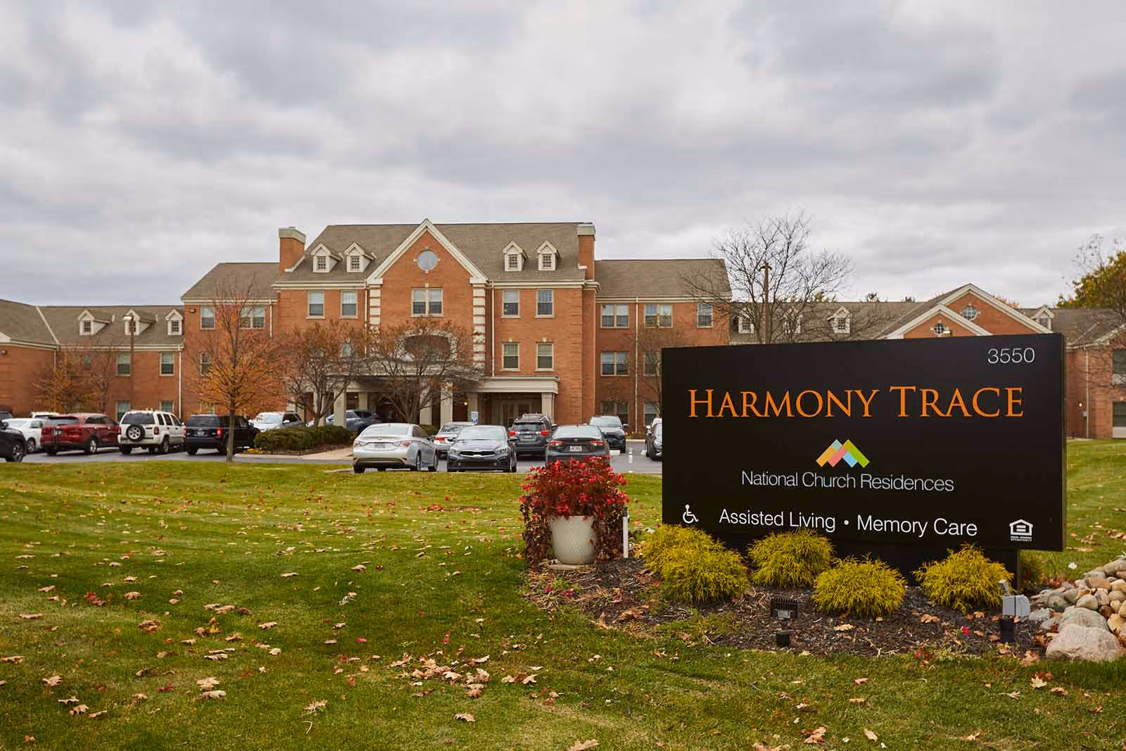 Exterior view of Harmony Trace senior living facility showing a large brick building with multiple windows and a parking lot with several cars. In the foreground, there is a black sign with orange and white text that reads 'Harmony Trace, National Church Residences, Assisted Living, Memory Care' surrounded by green grass and landscaping with bushes and flowers.