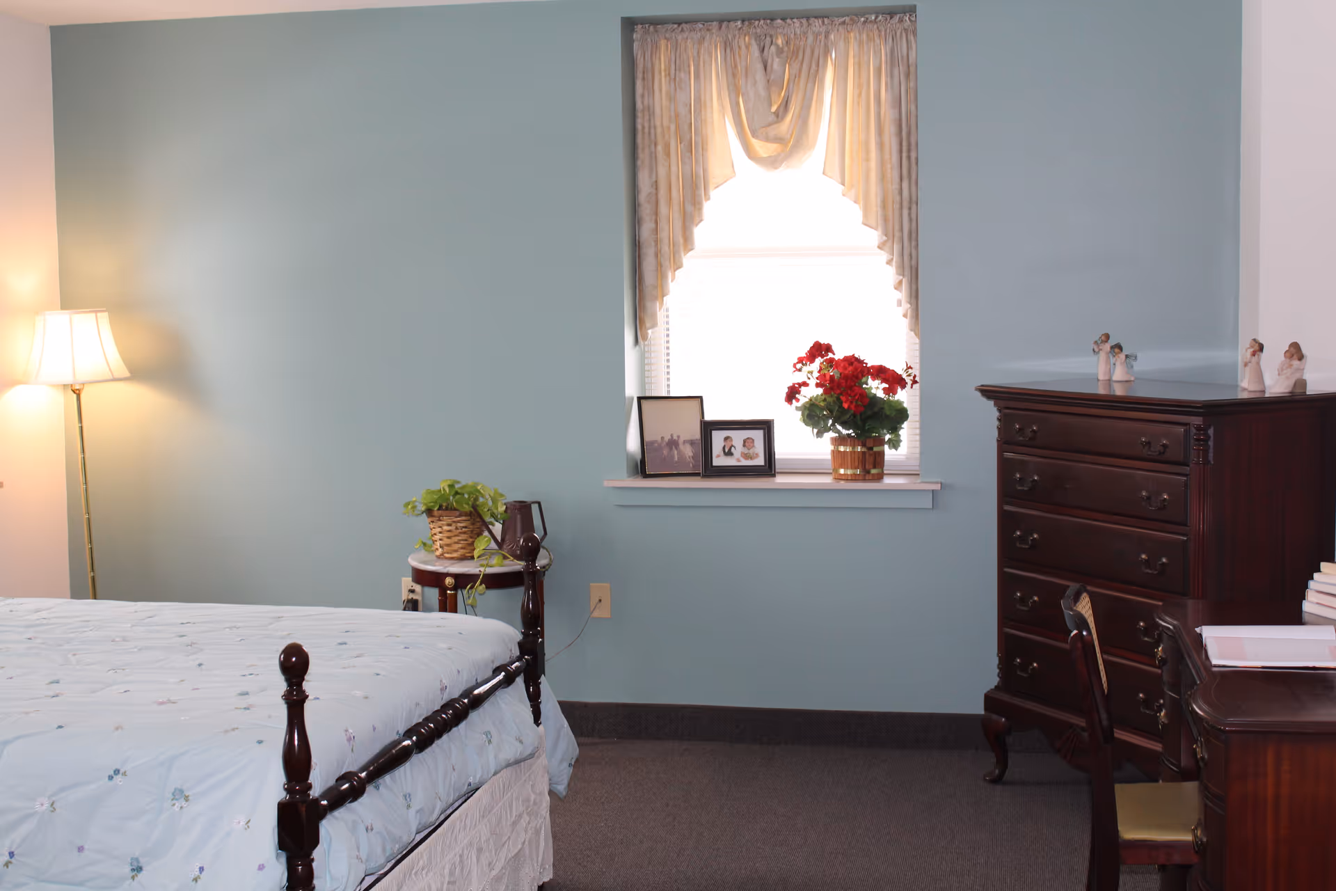 A bedroom with light blue walls featuring a bed with a light blue floral bedspread, a wooden nightstand with a potted plant and a lamp, a window with beige curtains, framed photos, a red flowering plant on the windowsill, a dark wooden dresser with figurines on top, and a wooden desk with a chair and some books.
