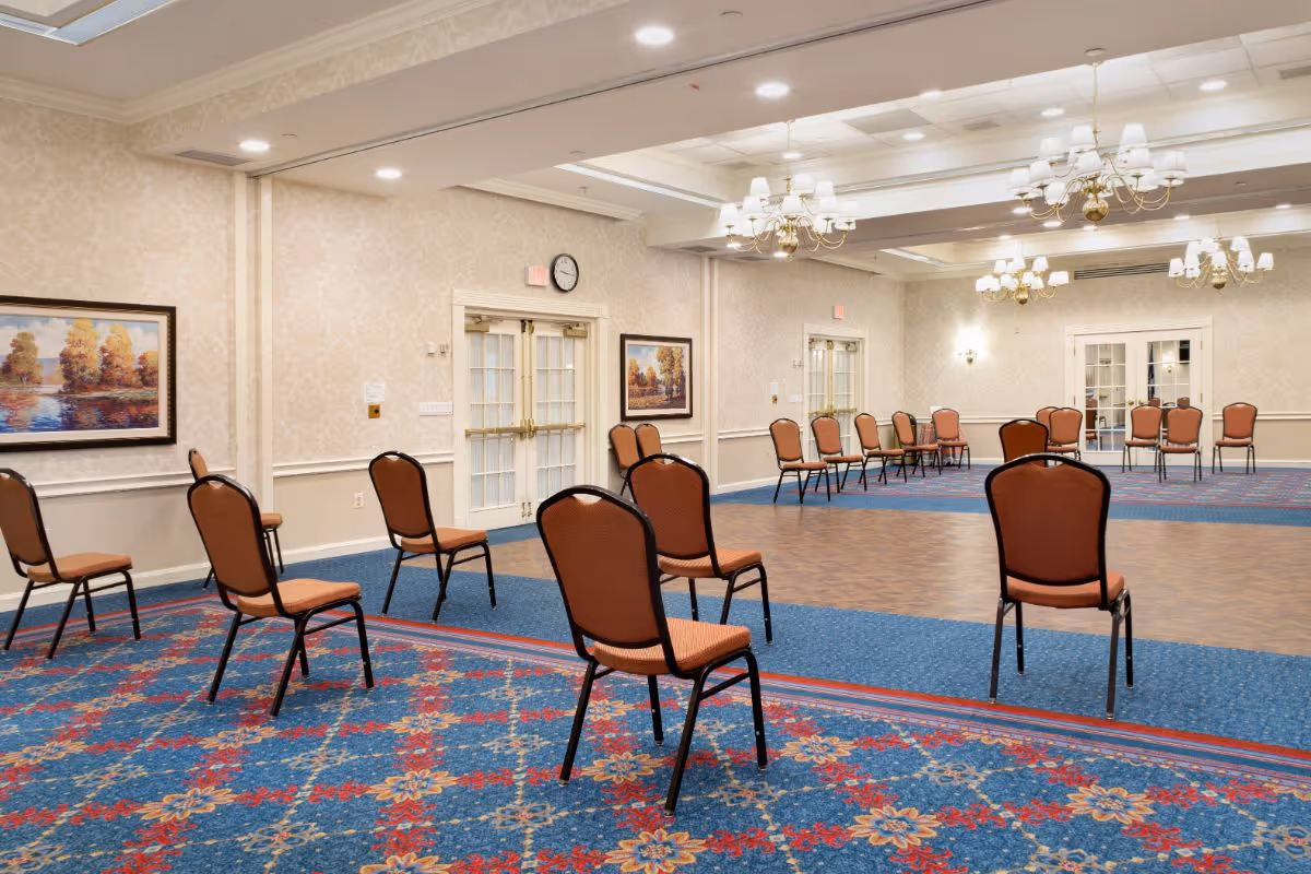 A large, well-lit meeting or event room with multiple brown cushioned chairs spaced apart on a patterned blue and red carpet. The room has beige wallpaper, framed landscape paintings on the walls, chandeliers hanging from the ceiling, and double glass doors on two sides.