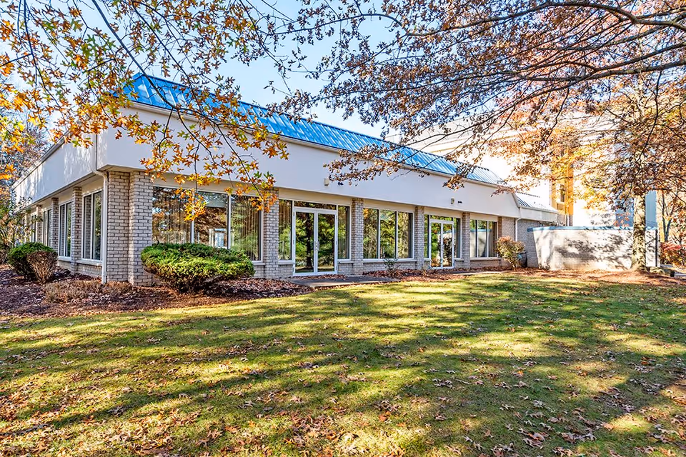 Exterior view of a single-story senior living facility building with large windows and a blue metal roof. The building is surrounded by a grassy lawn with scattered fallen leaves and some bushes. Trees with autumn-colored leaves frame the scene under a clear blue sky.
