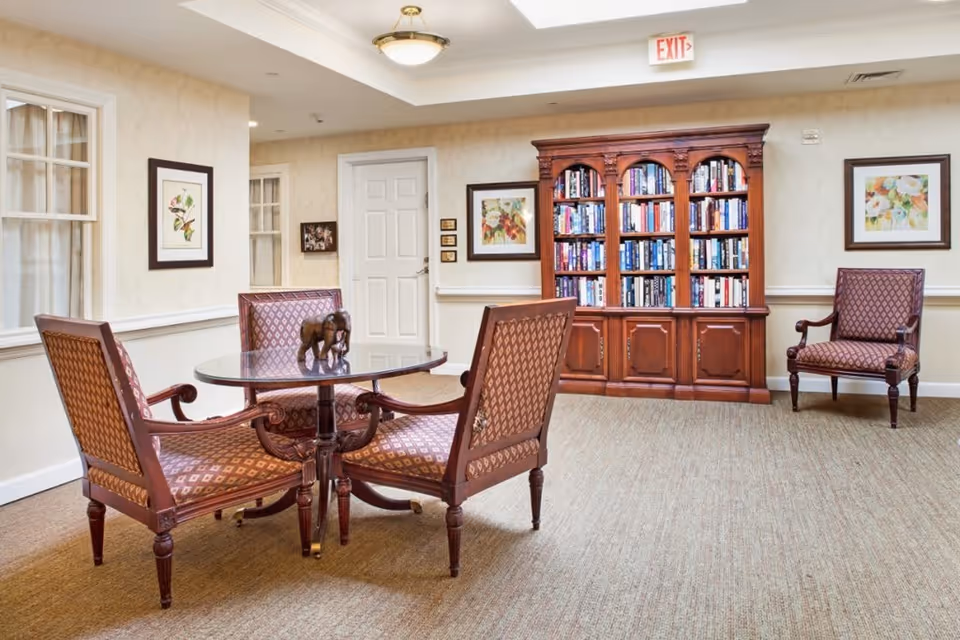 A cozy interior room with a round glass-top table surrounded by four upholstered wooden chairs. Behind the table is a large wooden bookshelf filled with books. The walls are decorated with framed floral artwork, and there is an exit sign above a white door in the background. The room has beige carpeting and soft lighting from a ceiling fixture.