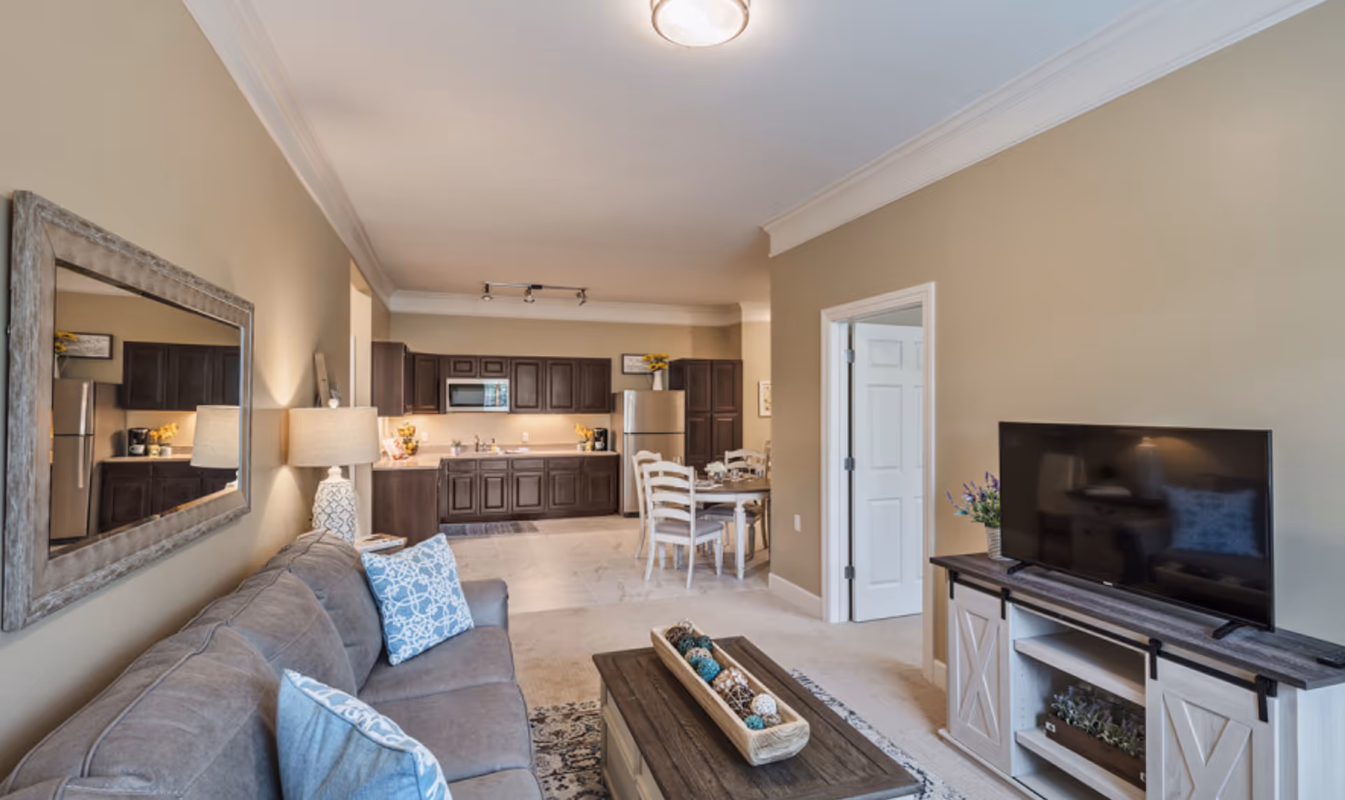 A cozy senior living apartment interior featuring a gray sofa with blue patterned pillows, a wooden coffee table with decorative items, a large wall mirror, a flat-screen TV on a white and wood TV stand, and an open kitchen and dining area with dark wood cabinets, stainless steel appliances, and a dining table with chairs.