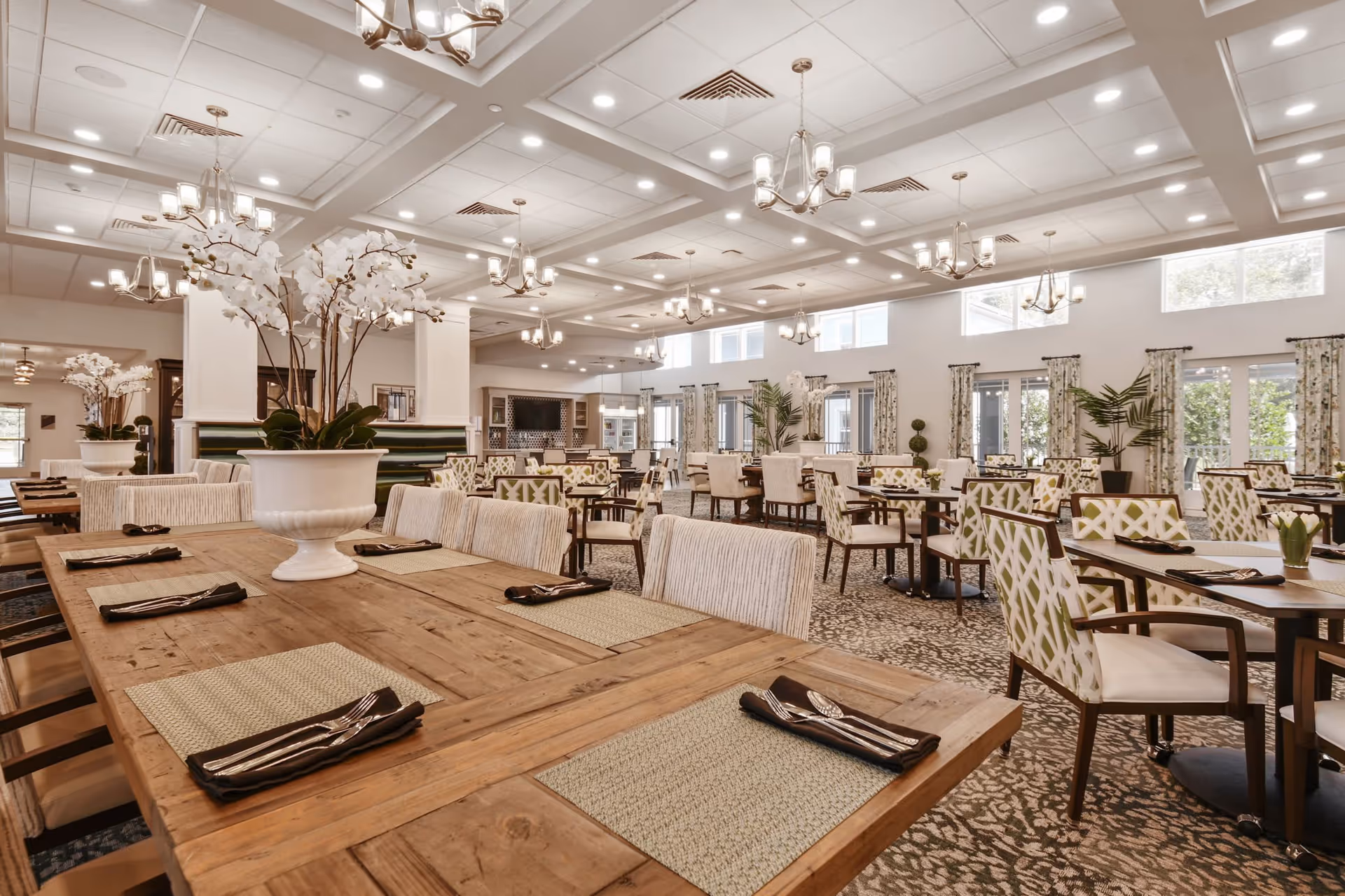A spacious and well-lit dining room with multiple wooden tables set with placemats, napkins, and silverware. The room features comfortable upholstered chairs with patterned fabric, large windows with curtains, decorative plants, and elegant chandeliers hanging from a coffered ceiling.
