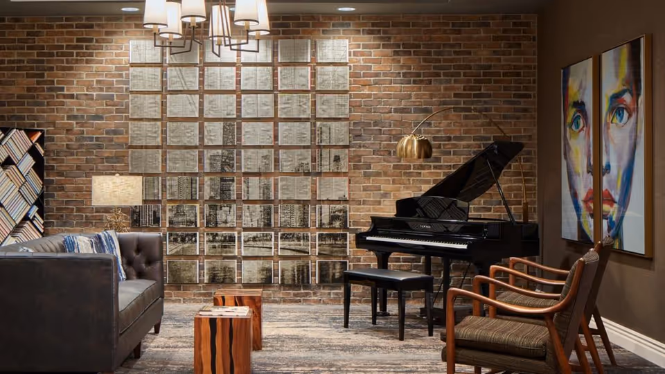 A cozy living room with a dark leather sofa on the left, two wooden stools in the center, and two wooden armchairs with cushions on the right. A black grand piano with a matching bench is positioned against a brick wall, which is decorated with a grid of framed artwork. A modern floor lamp with a gold shade arches over the piano. On the right wall, there is a colorful abstract portrait painting. The room is softly lit by a ceiling light fixture and a table lamp on the left side.