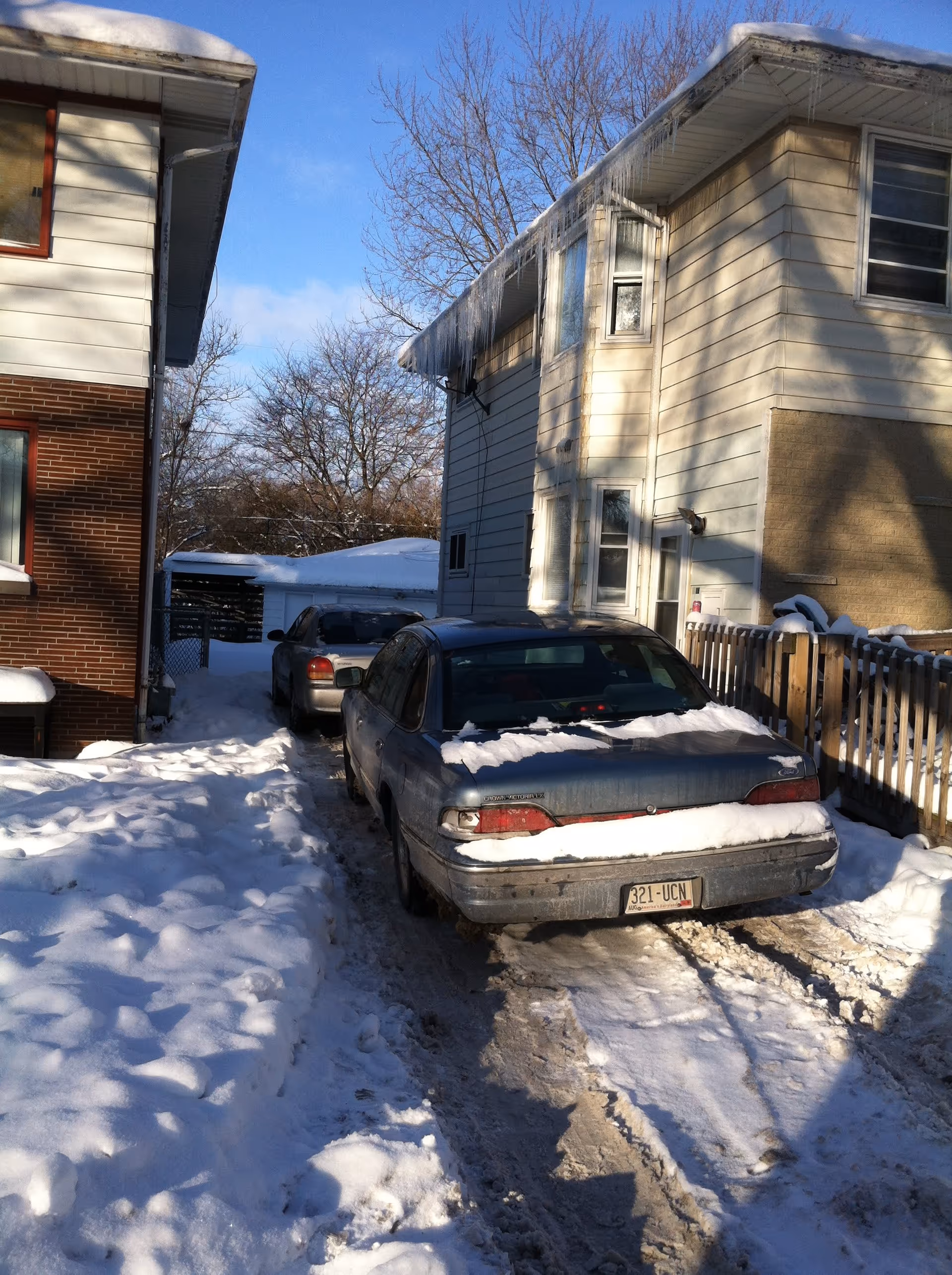 Snow-covered driveway between two houses with two parked cars, one closer to the camera partially covered in snow. Icicles hang from the roof of the house on the right. Bare trees and a clear blue sky are visible in the background.