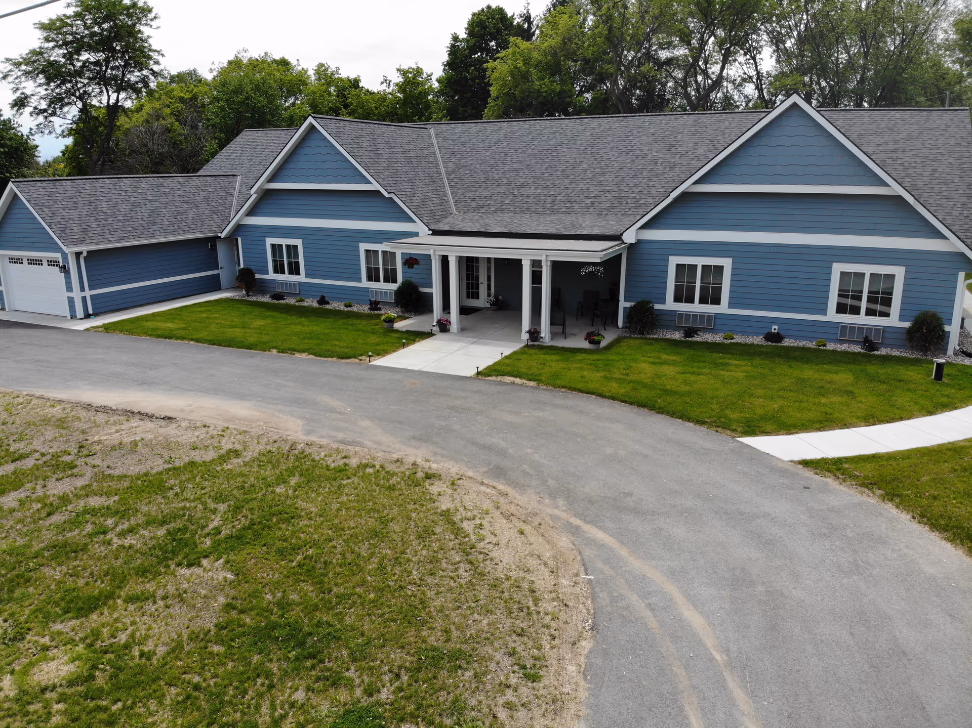A single-story blue building with white trim and a gray roof, surrounded by green grass and a paved driveway. The building has a covered entrance with a small porch area and several windows. Trees are visible in the background.