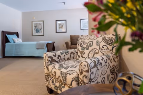 A cozy senior living room with a patterned armchair in the foreground, a brown sofa, and a single bed with light blue bedding against the wall. Three framed pictures hang on the beige wall behind the sofa and bed. A vase with colorful flowers is partially visible on the right side.