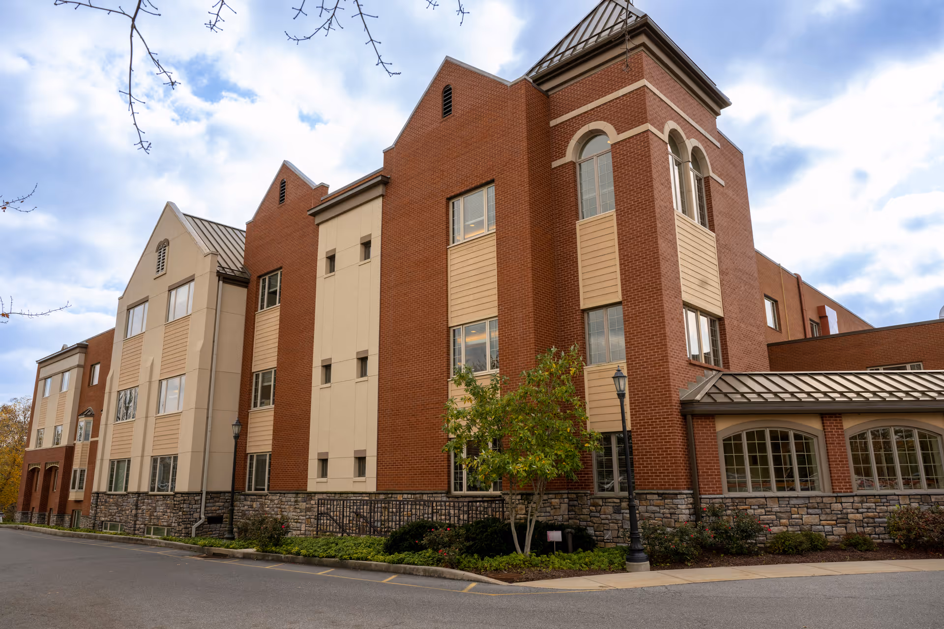 Exterior view of a multi-story senior living facility building with brick and beige paneling, stone accents at the base, several windows, a small tree, and a street lamp in front under a partly cloudy sky.