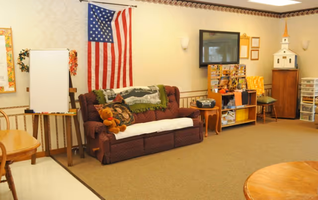 A cozy common room with a maroon couch adorned with a teddy bear and pillows, an American flag hanging on the wall, a whiteboard on an easel, a wooden table and chairs, a small bookshelf with various items, a wall-mounted TV, and a model of a white church on a cabinet.