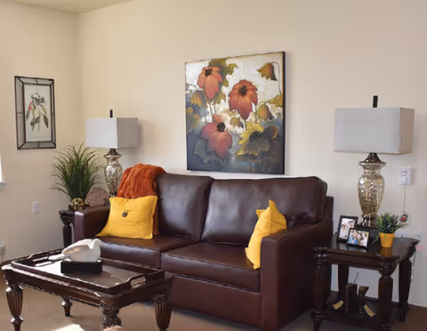 A cozy living room with a dark brown leather sofa adorned with two yellow pillows and an orange throw blanket. There are two matching glass table lamps with beige shades on dark wooden side tables on either side of the sofa. A coffee table with decorative items is in front of the sofa. The walls are light-colored and decorated with a large floral painting above the sofa and a framed botanical print on the adjacent wall.