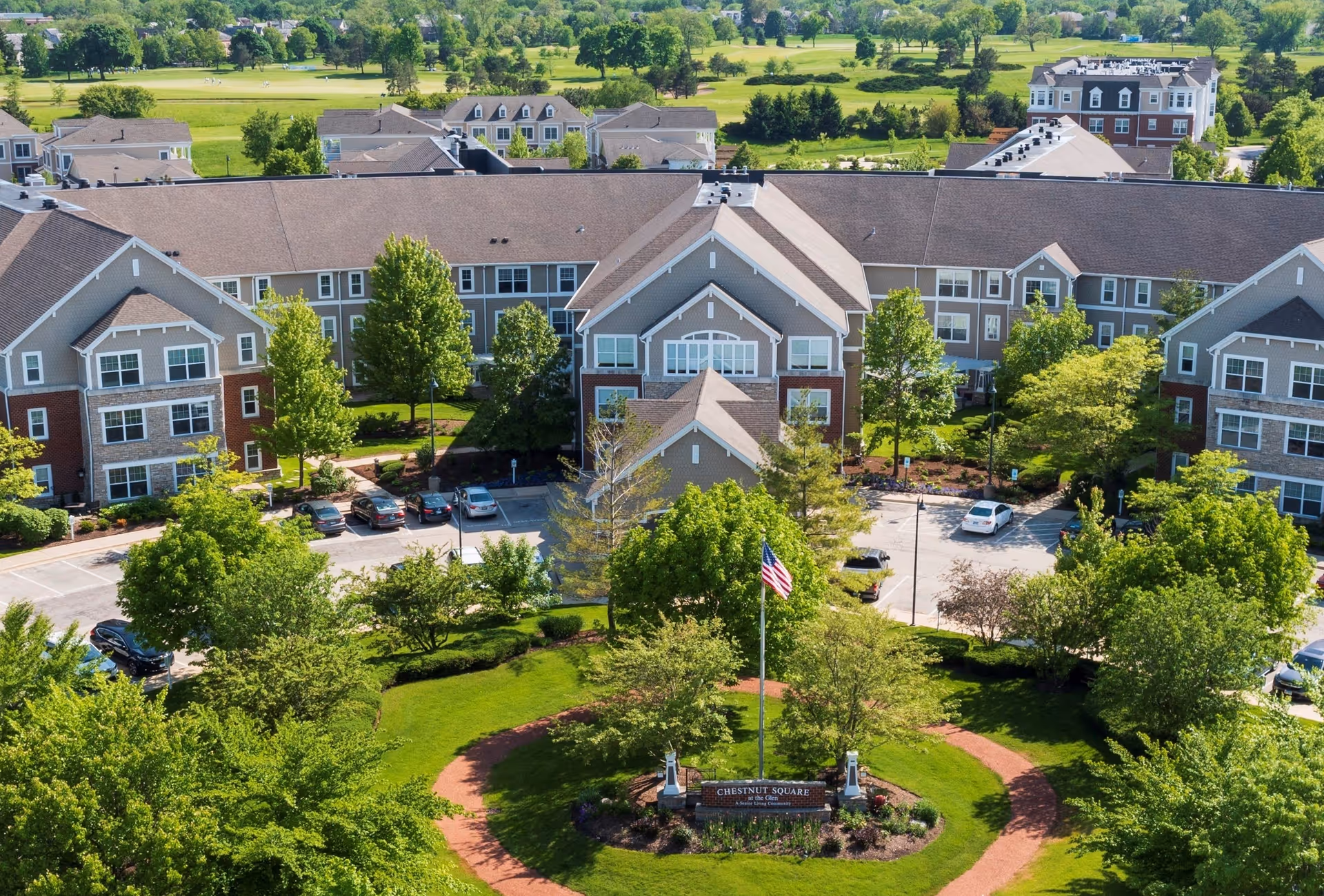 Aerial view of Chestnut Square at The Glen senior living facility showing multiple connected residential buildings surrounded by green trees and landscaped gardens. A circular pathway encircles a flagpole with an American flag and a sign that reads 'Chestnut Square at The Glen'. Cars are parked in the parking lot near the buildings.
