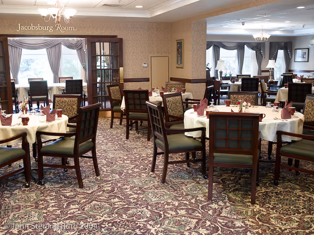 A spacious dining room with round tables covered in white tablecloths, each set with folded napkins, glassware, and floral centerpieces. The room features patterned carpet, wooden chairs with green cushions, and large windows with curtains allowing natural light. A sign reading 'Jacobsburg Room' is visible above a doorway with glass-paneled double doors.