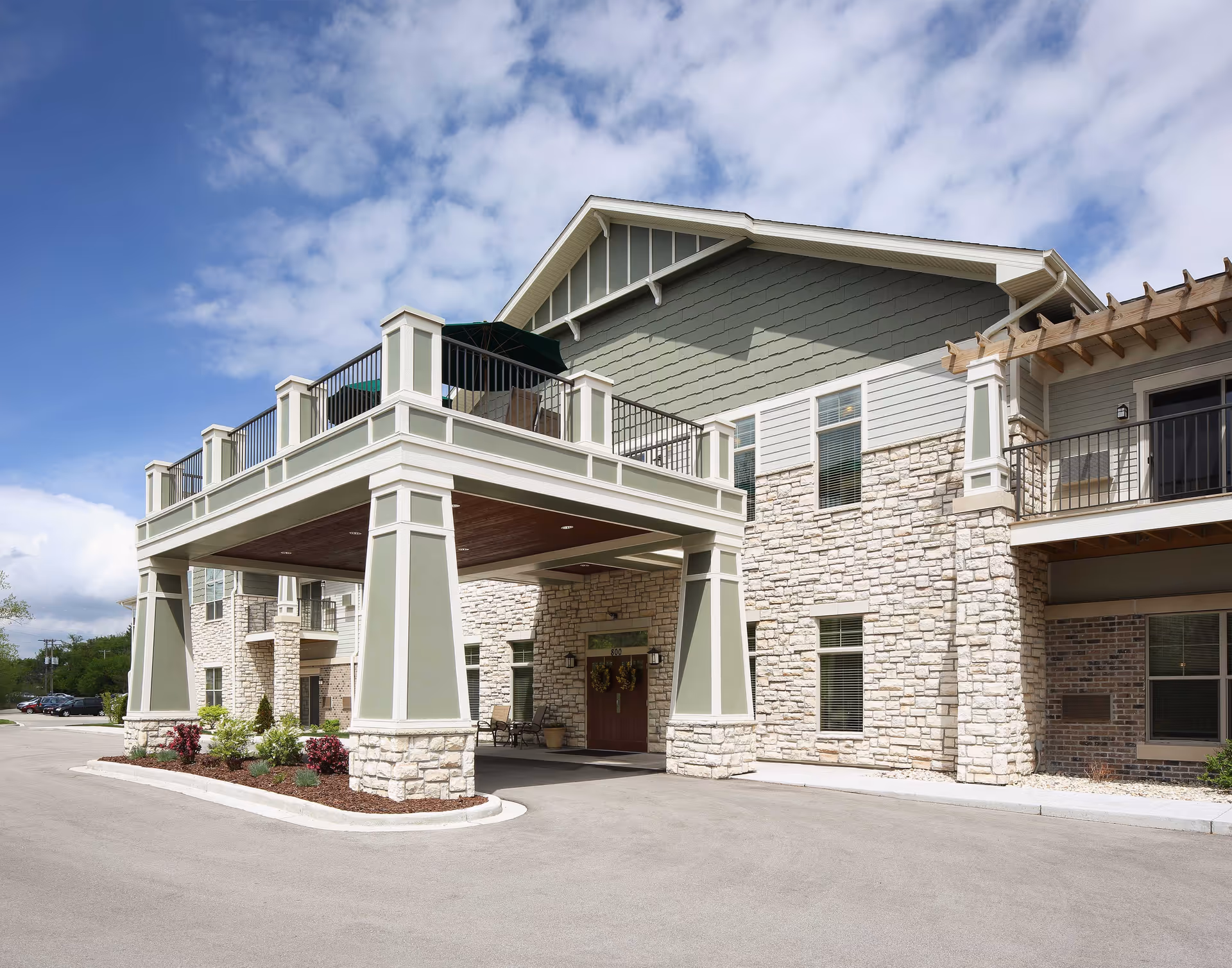 Exterior view of a senior living facility building with stone and siding facade, featuring a covered entrance supported by large pillars and a balcony above. The sky is partly cloudy and there are some plants and shrubs near the entrance.