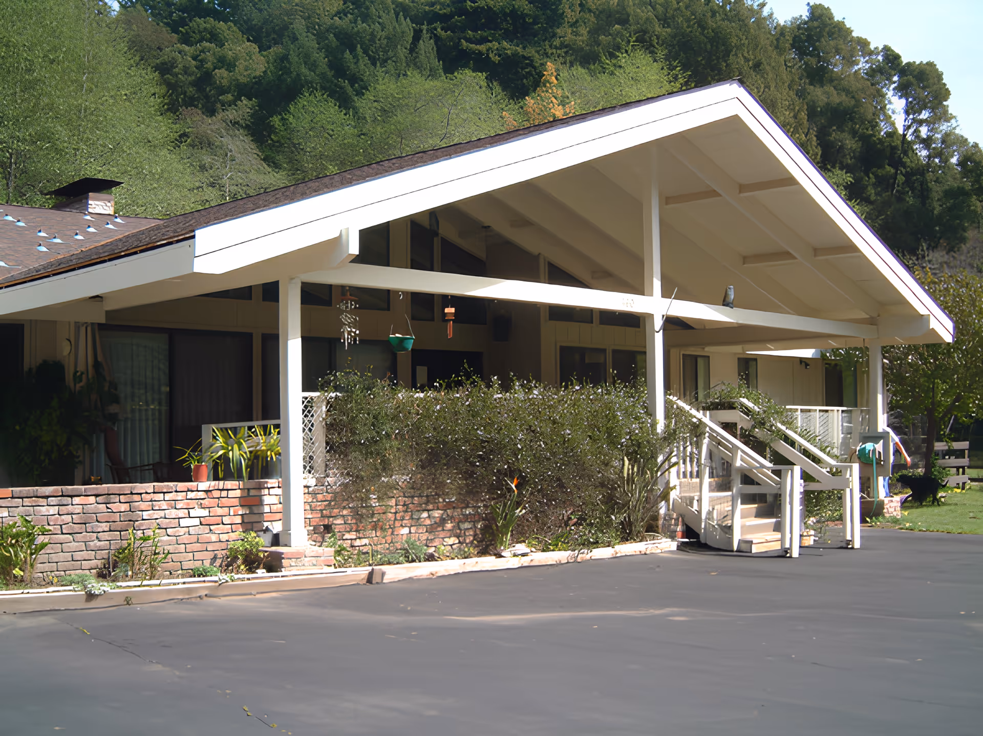 Exterior view of a single-story building with a large white slanted roof, brick planter boxes with green plants, and a paved driveway in front. The building is surrounded by trees and greenery.