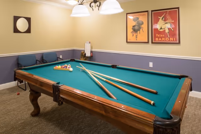 A room with a pool table set up for a game, featuring pool cues resting on the table and a rack of billiard balls. The room has beige and blue walls with two framed vintage posters and a round mirror. There are three blue chairs against the wall and a ceiling light fixture above the pool table.