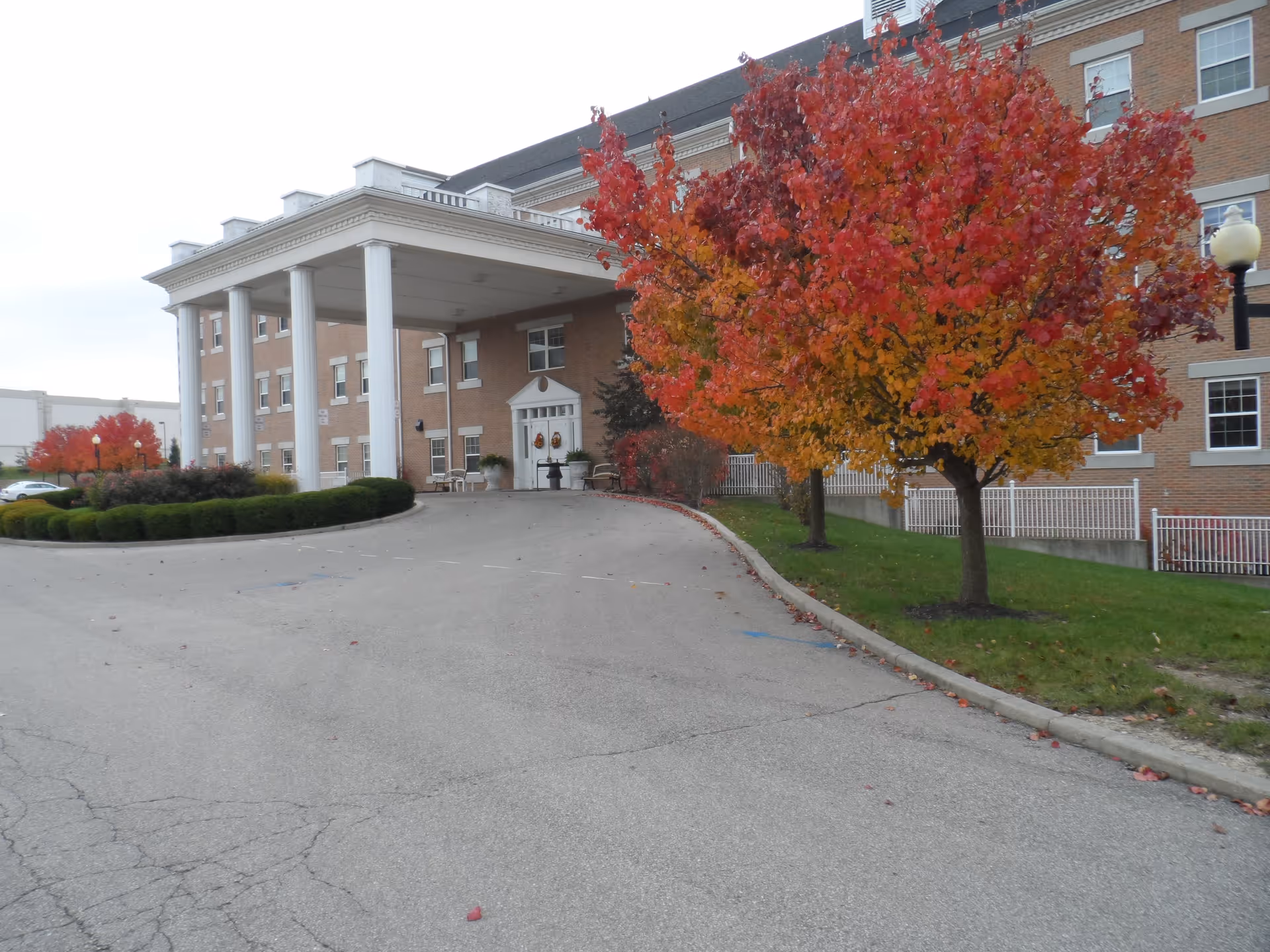 Exterior view of a multi-story brick building with a large covered entrance supported by white columns. There are vibrant autumn trees with red and orange leaves along the driveway leading to the entrance.