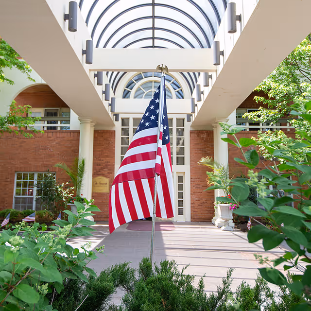 Entrance of The Gatesworth facility with a glass arched roof, brick walls, white columns, and an American flag in the foreground surrounded by greenery.