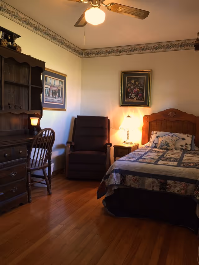 A cozy bedroom with a wooden bed featuring a patchwork quilt and decorative pillows. Next to the bed is a wooden nightstand with a lit table lamp. A dark upholstered recliner chair is positioned against the wall, and a wooden desk with a matching chair is on the left side. The room has hardwood floors, floral wallpaper border near the ceiling, and framed artwork on the walls. A ceiling fan with a light is visible above.