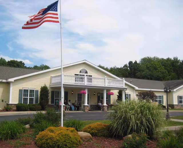 Exterior view of a single-story yellow building with white trim, featuring a covered entrance with columns and a balcony above. There are several people sitting on the porch, an American flag on a flagpole in the front, and landscaped bushes and plants in the foreground under a partly cloudy sky.
