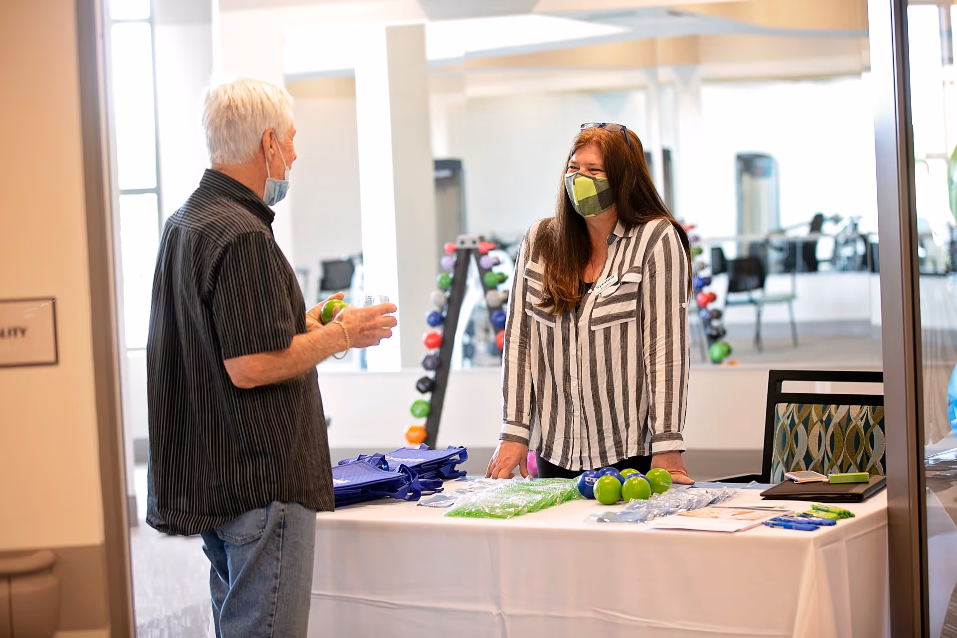 An elderly man wearing a face mask talks to a woman behind a table covered with promotional items including stress balls and pens. They are indoors in a bright room with exercise equipment and mirrors in the background.