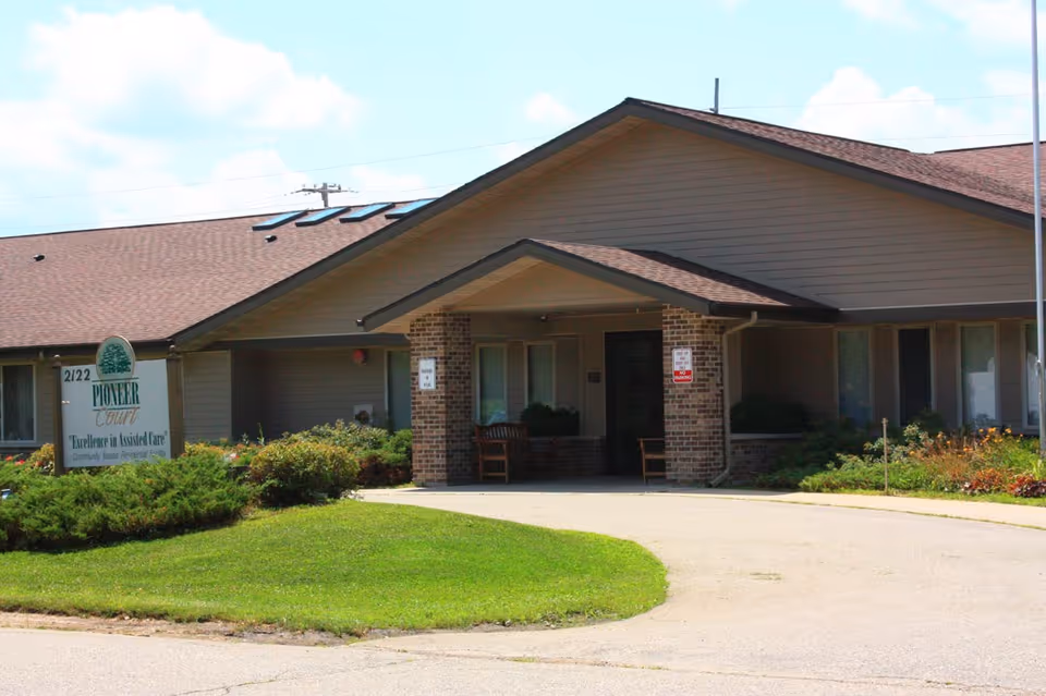 Exterior view of a single-story assisted living facility building with a covered entrance supported by brick pillars. There is a driveway leading to the entrance, green grass, and bushes around the building. A sign near the entrance reads 'Pioneer Court Excellence in Assisted Care'.