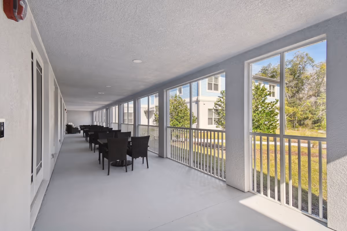 A long covered outdoor patio area with multiple black wicker chairs and tables arranged in pairs along the left side. The right side has large screened openings with white railings, allowing a view of green trees and a neighboring building. The floor and ceiling are light-colored, and the space appears bright and airy.