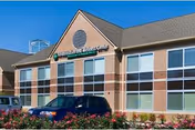 Exterior view of a brick and glass building with a sign that reads 'Fraser Villa Wellness Center'. There are several windows and a parking lot in front with cars parked. Flowering bushes line the edge of the parking area.