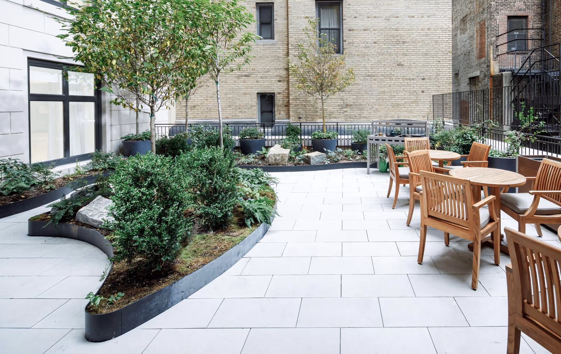 Outdoor patio area with white tiled flooring, wooden tables and chairs with cushions, and a garden bed containing small trees and shrubs. The patio is enclosed by a black metal fence and surrounded by brick and stone building walls.