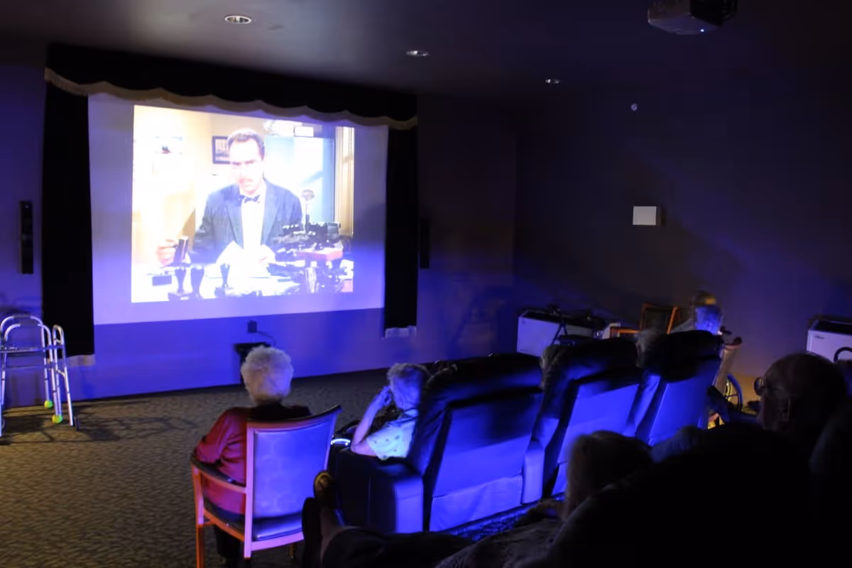 A group of elderly people seated in a dimly lit room watching a movie projected on a large screen. Some are sitting in recliners while one person is in a chair. A walker is visible on the left side of the room.