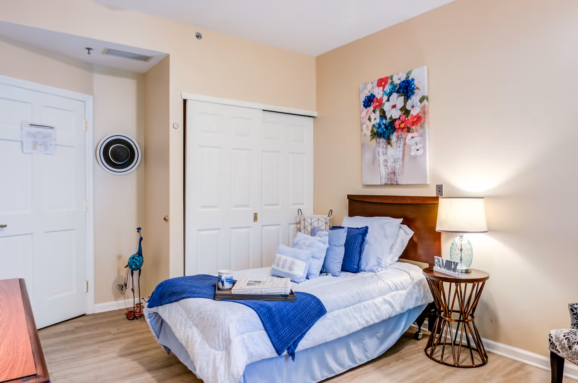 A cozy bedroom with a single bed made up with white and blue bedding, multiple pillows, and a blue throw blanket. A wooden headboard is against the beige wall, above which hangs a colorful floral painting. To the right of the bed is a round wooden side table with a lamp and a small decorative sign that says 'FAMILY'. There is a white closet with sliding doors and a white door on the left side of the room. The floor is light wood, and the walls are painted beige.